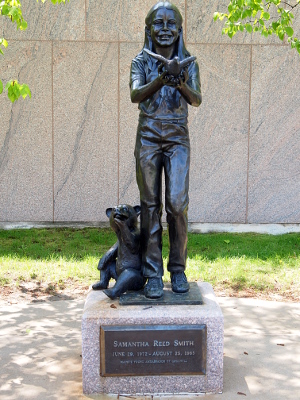 [Metal statue of a young girl standing with her hands together holding a dove which is taking flight. Beside her sits a small bear with one paw covering its mouth. The statue sits on a base with a nameplate attached to the front describing it.]